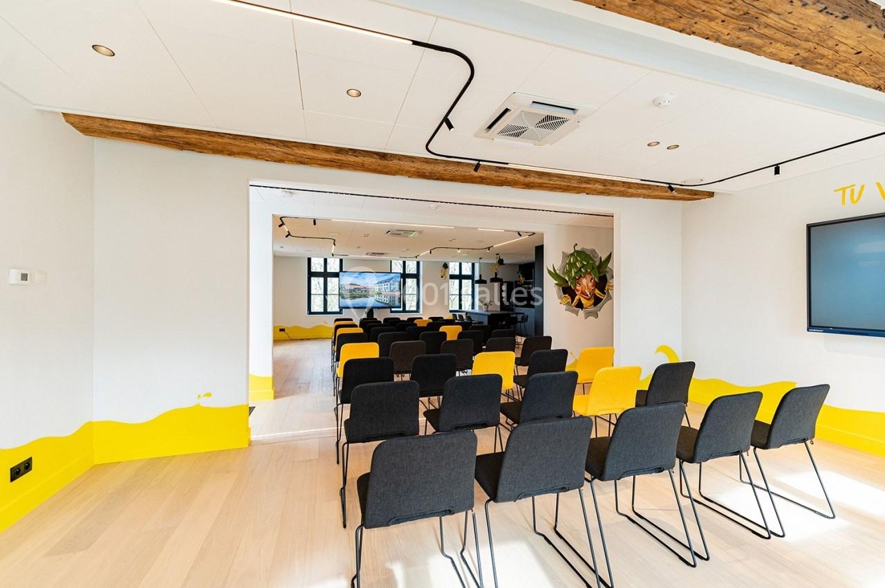 Salle de réunion lumineuse avec rangées de chaises noires, écran mural et poutres apparentes.