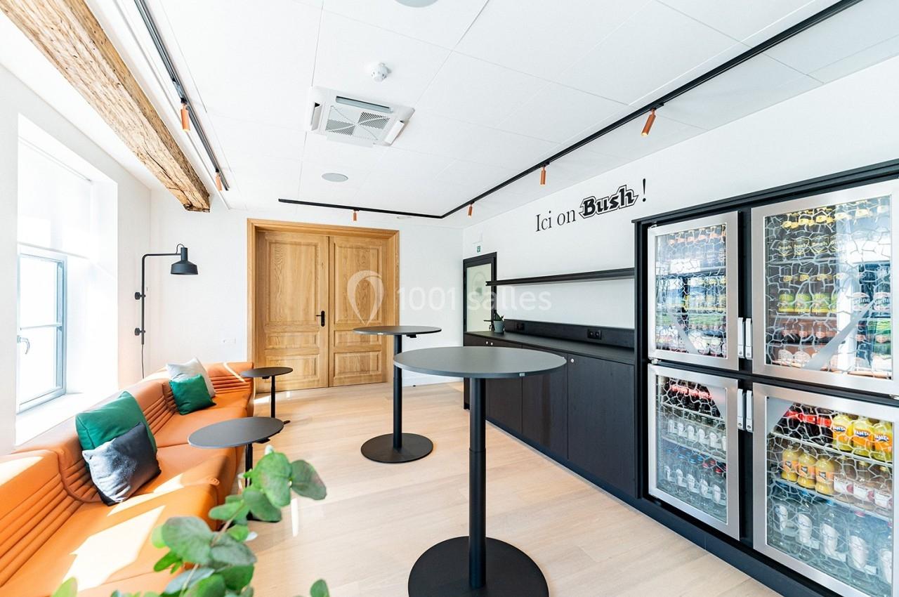 Espace lumineux avec tables hautes, banquette orange, plantes et réfrigérateurs remplis de boissons.