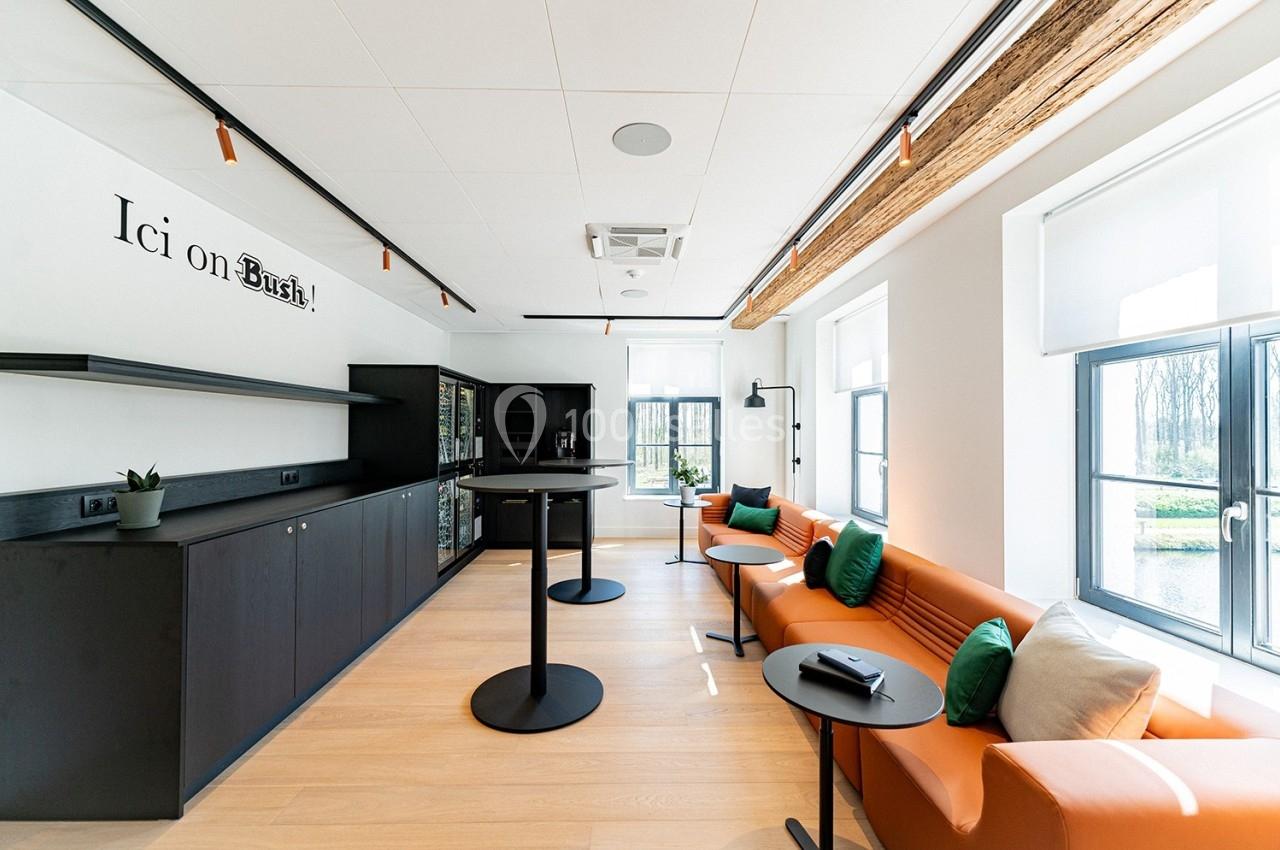 Espace lumineux avec canapé orange, tables hautes noires, étagères et grandes fenêtres donnant sur l'extérieur.