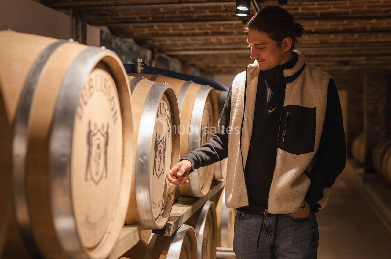 Un homme observe attentivement des fûts en bois alignés dans une cave voûtée.