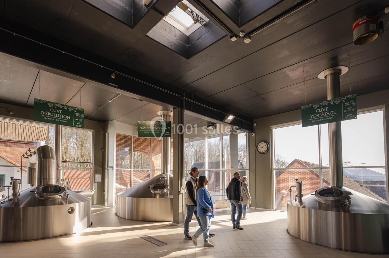 Visiteurs marchant dans une brasserie lumineuse avec cuves en acier inoxydable et panneaux explicatifs suspendus.