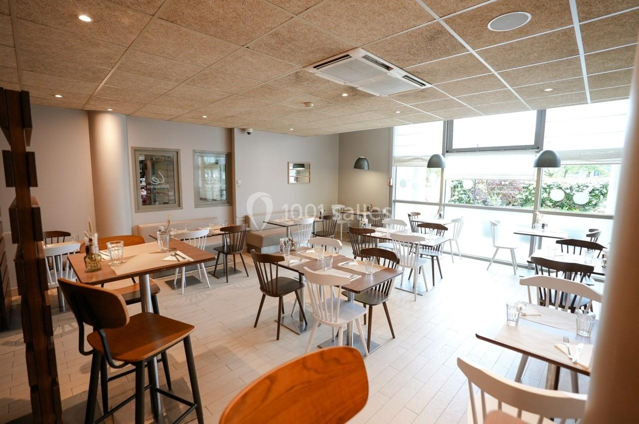 Salle de restaurant lumineuse avec tables en bois clair, chaises variées et grandes fenêtres donnant sur l'extérieur.