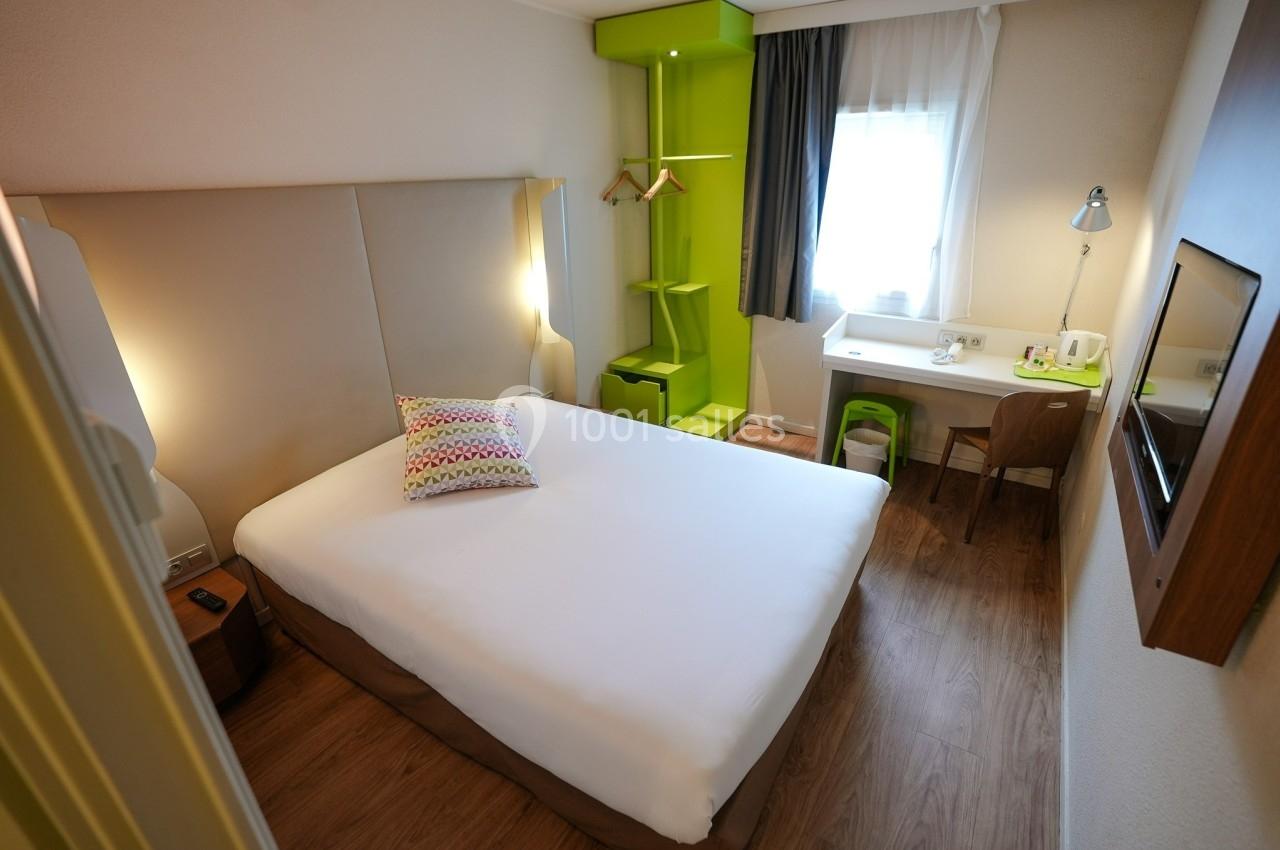 Chambre d'hôtel lumineuse avec lit double, bureau, chaise, étagère verte et fenêtre laissant entrer la lumière naturelle.