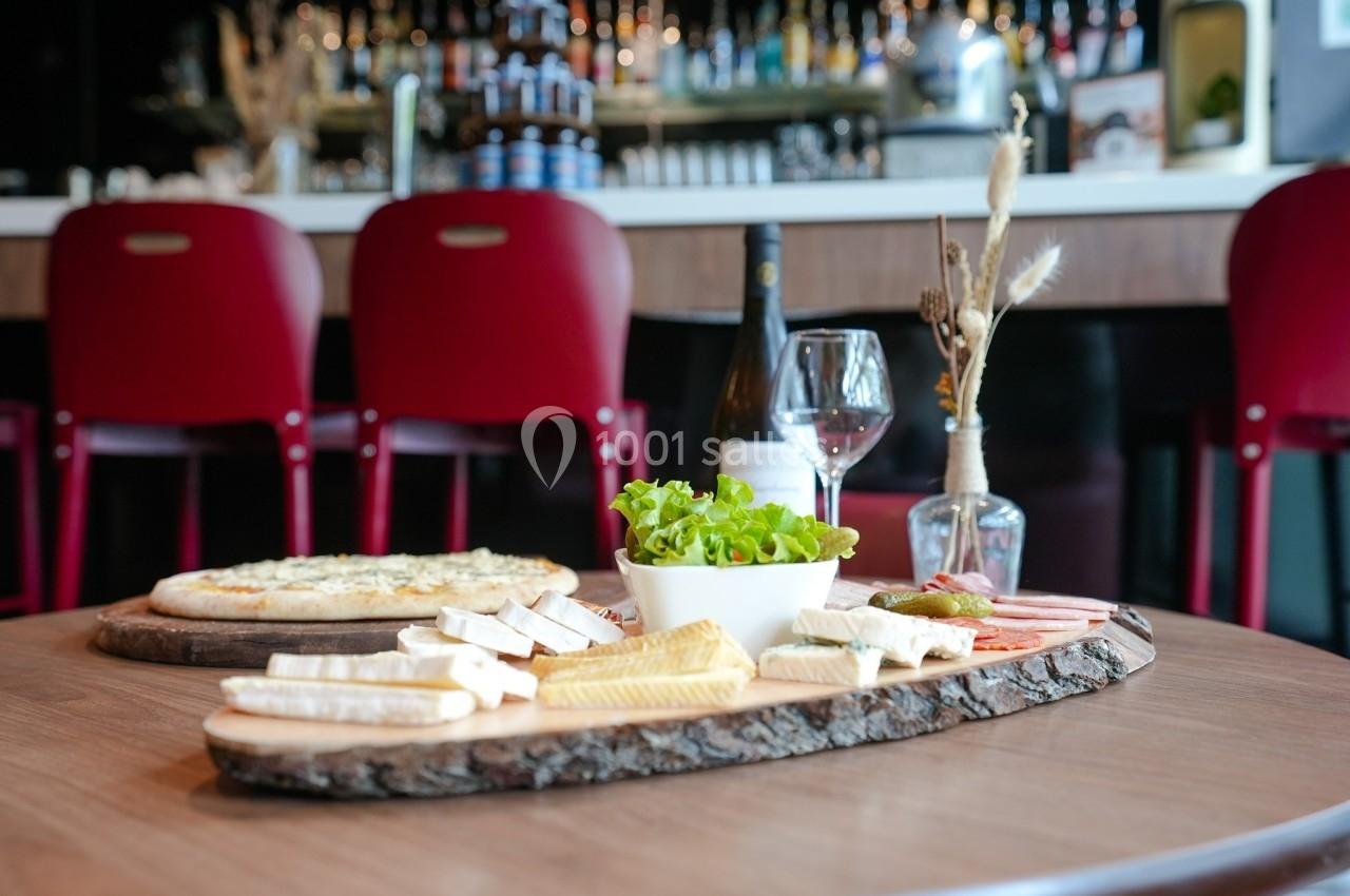 Plateau de fromages et charcuteries avec salade, posé sur une table en bois, devant un bar avec des chaises rouges.