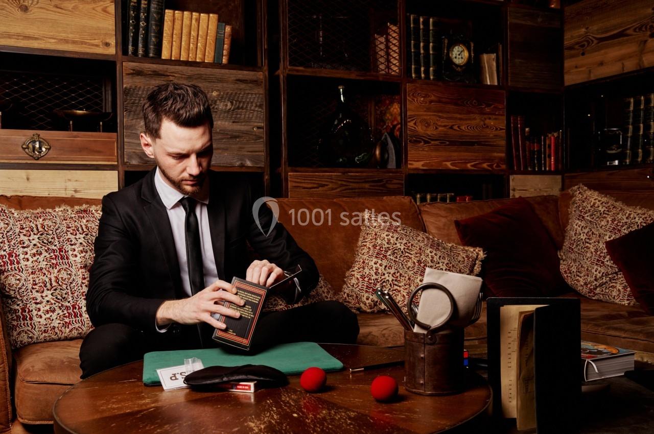 Un homme en costume assis sur un canapé, manipulant un livre dans un salon décoré de bois et d'étagères remplies de livres.