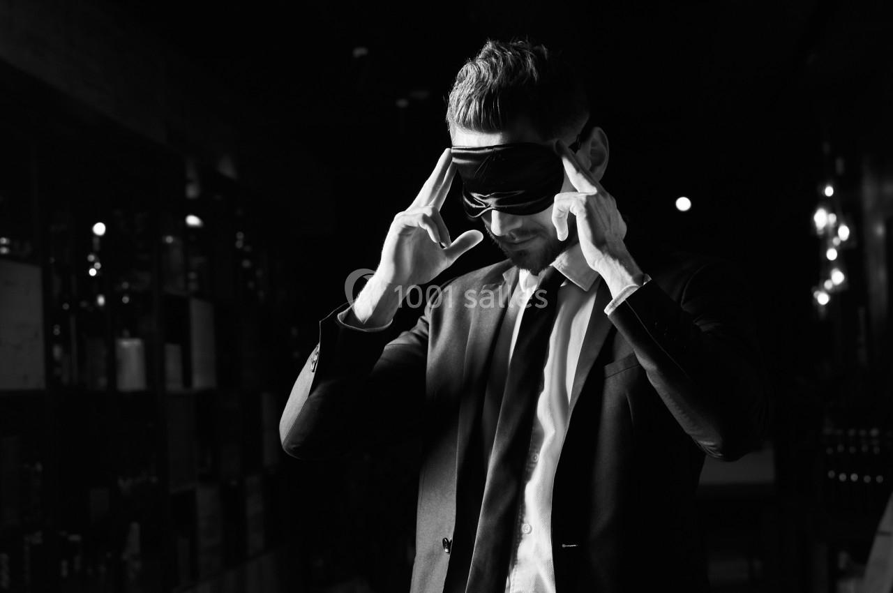 Homme en costume ajustant un bandeau noir sur ses yeux dans un environnement sombre.