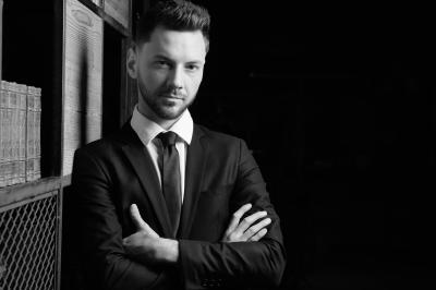 Un homme en costume, les mains levées, portant un masque noir sur les yeux dans un environnement sombre.