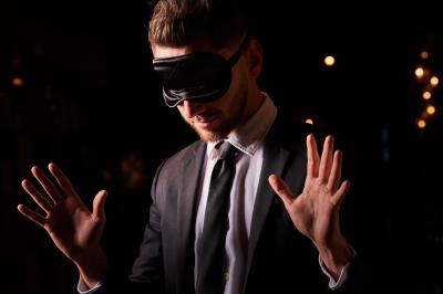 Un homme en costume, les mains levées, portant un masque noir sur les yeux dans un environnement sombre.