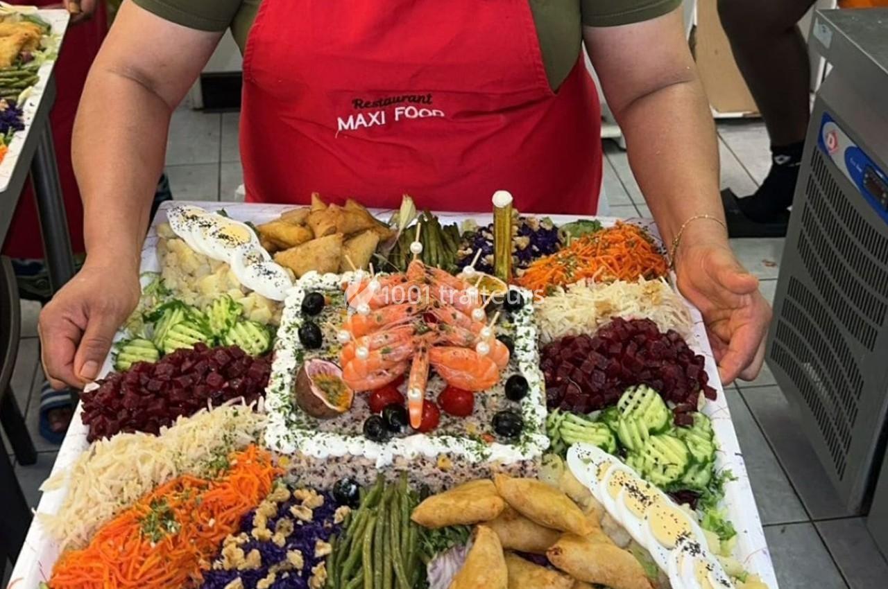Plateau garni de crudités, crevettes, samoussas et légumes variés, présenté par une personne en tablier rouge.