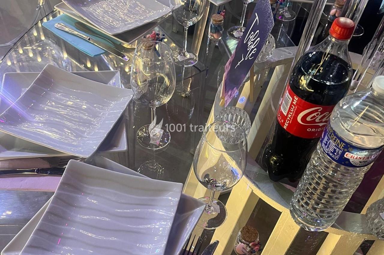 Table dressée avec des assiettes blanches, verres, bouteilles d'eau et de soda sur une surface réfléchissante.