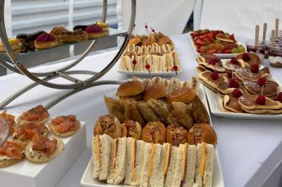 Buffet avec assortiments de sandwichs, samoussas, mini-burgers, canapés et coupes de fruits sur une table blanche.