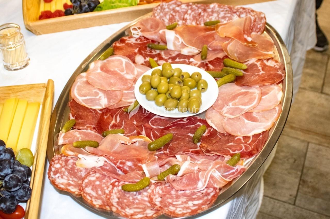 Plateau de charcuterie avec jambon, salami, cornichons et olives au centre, présenté sur une table blanche.