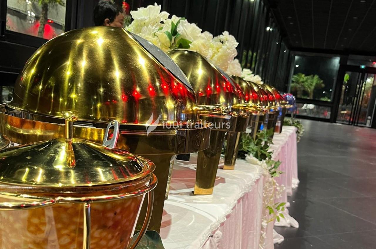 Rangée de plats en métal doré sur une table, dans une salle avec des baies vitrées et une décoration florale.