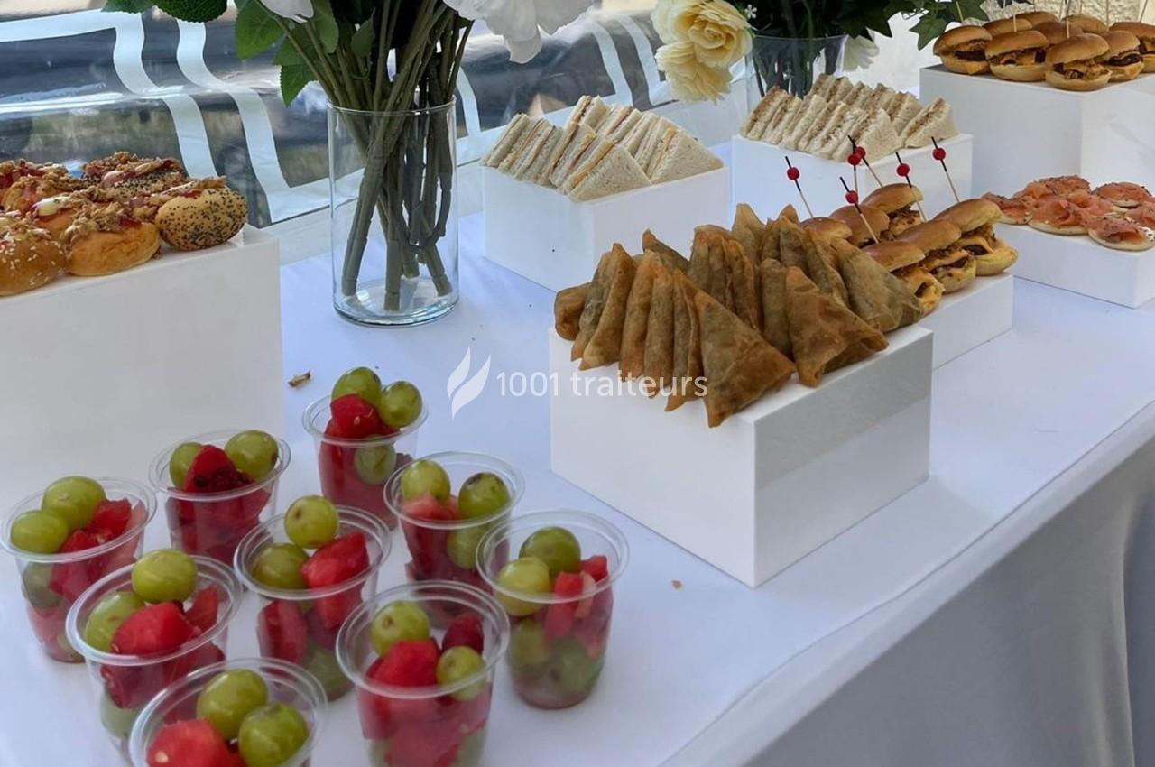 Buffet avec assortiments de sandwichs, samoussas, mini-burgers, canapés et coupes de fruits sur une table blanche.