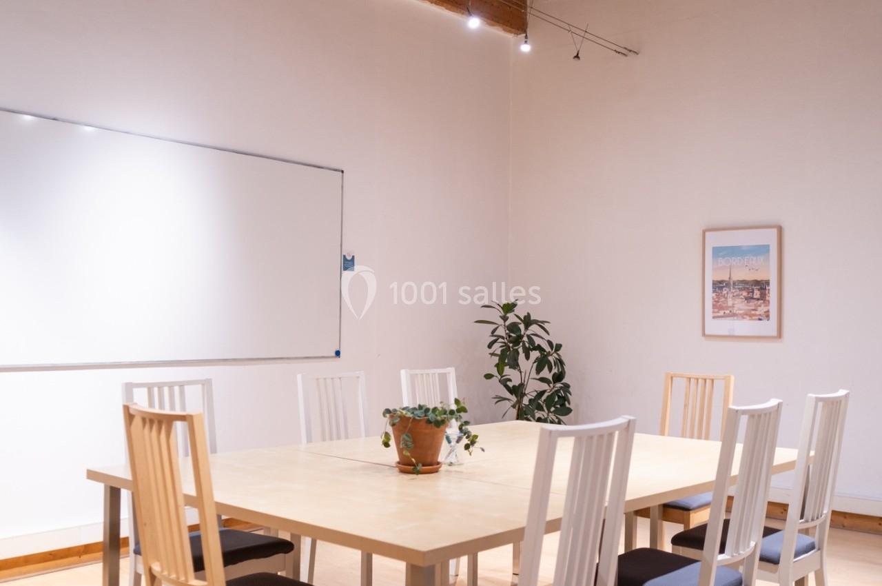 Salle de réunion lumineuse avec table en bois, chaises blanches, tableau blanc, plante et plafond en bois avec ventilateur.