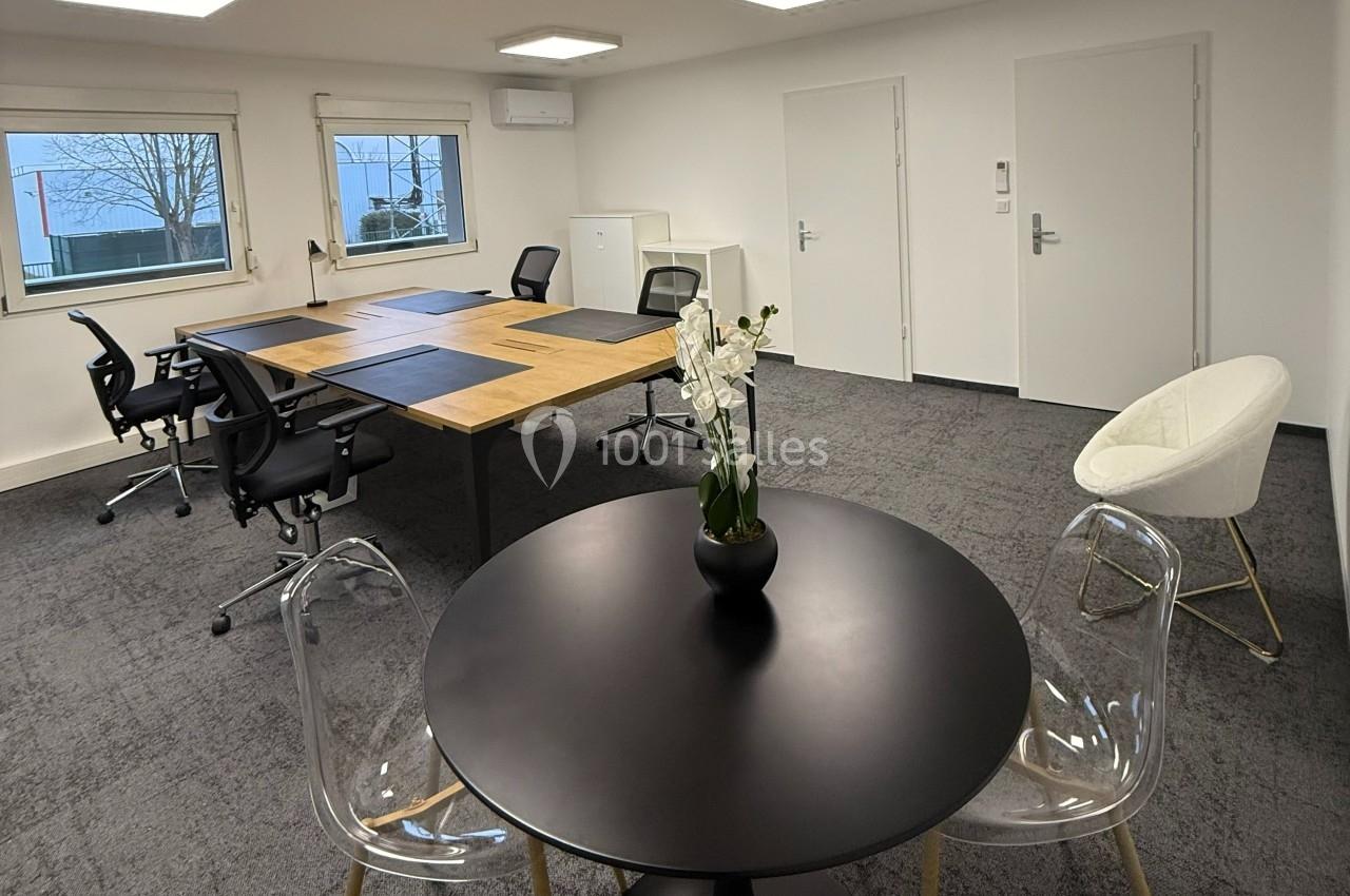 Salle de réunion moderne avec table ronde, chaises transparentes, bureau partagé et éclairage au plafond.