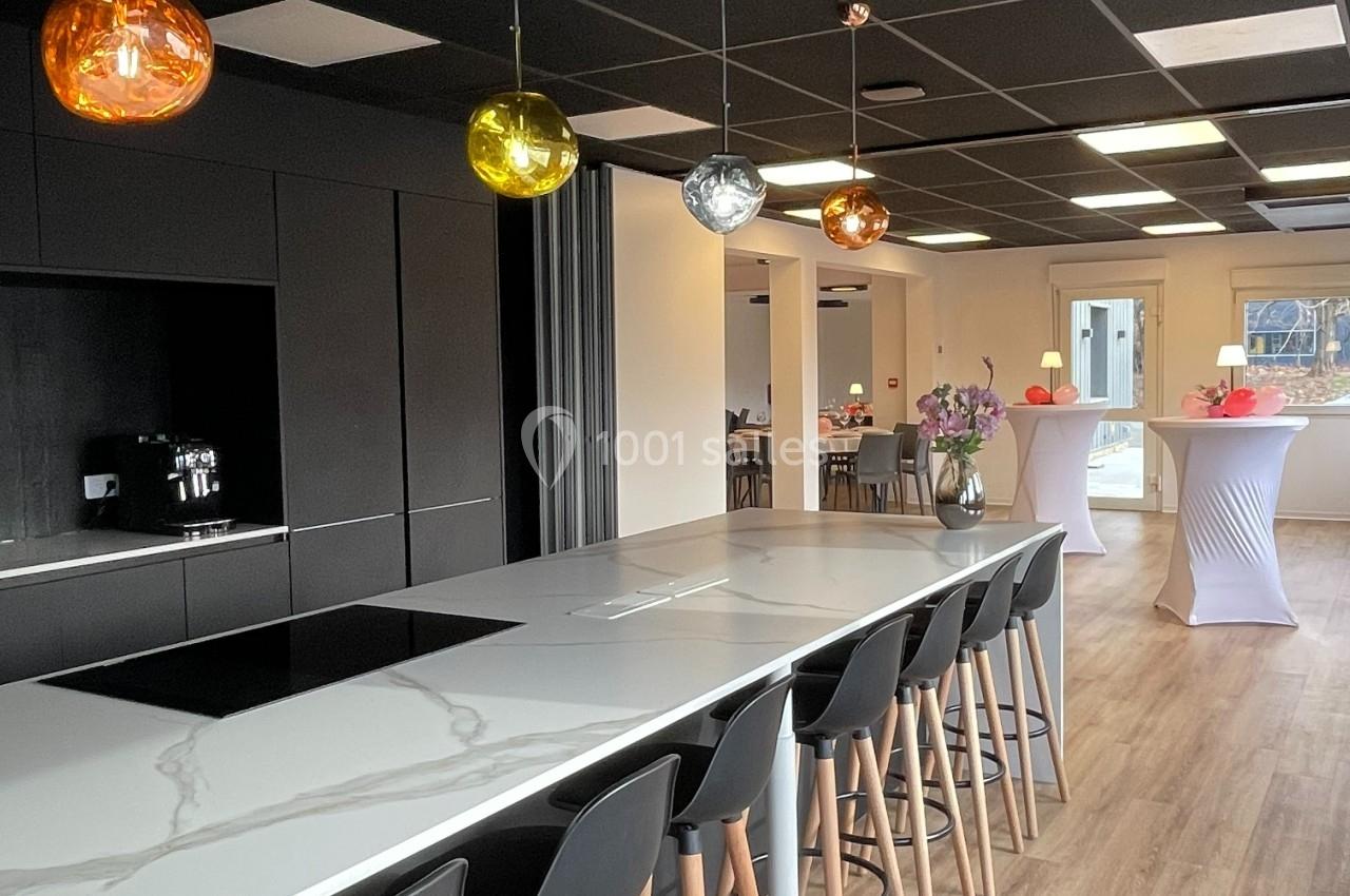 Cuisine moderne avec îlot central, tabourets noirs, suspensions colorées et vue sur une salle aménagée pour un événement.