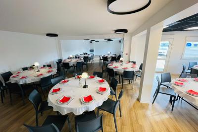 Salle de réception avec tables rondes dressées, nappes blanches, serviettes rouges et chaises grises sur parquet clair.