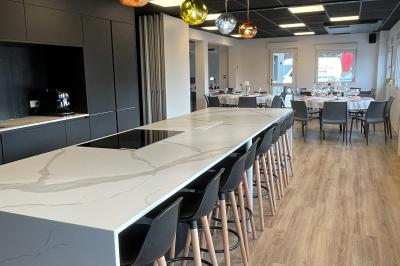 Salle de réception avec tables rondes dressées, nappes blanches, serviettes rouges et chaises grises sur parquet clair.