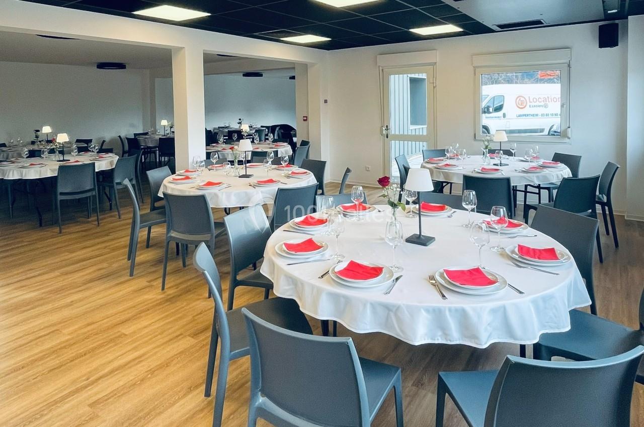 Salle de réception avec tables rondes dressées, nappes blanches, serviettes rouges et chaises grises sur parquet clair.