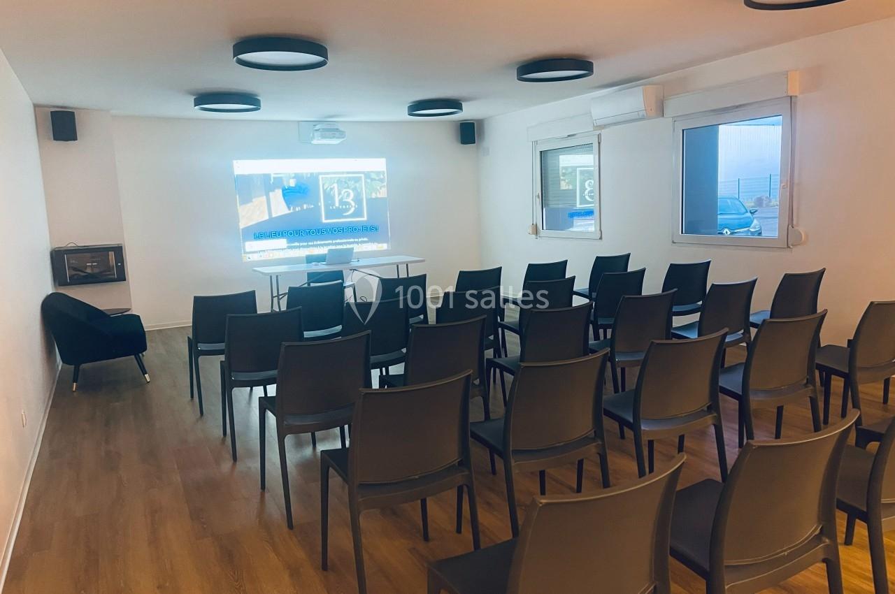 Salle de réunion lumineuse avec rangées de chaises noires, écran de projection allumé et parquet au sol.
