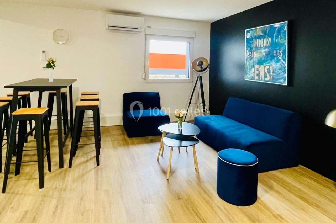 Salle moderne avec canapé bleu, tabourets hauts, tables noires, lampadaire et décoration murale sur fond sombre.