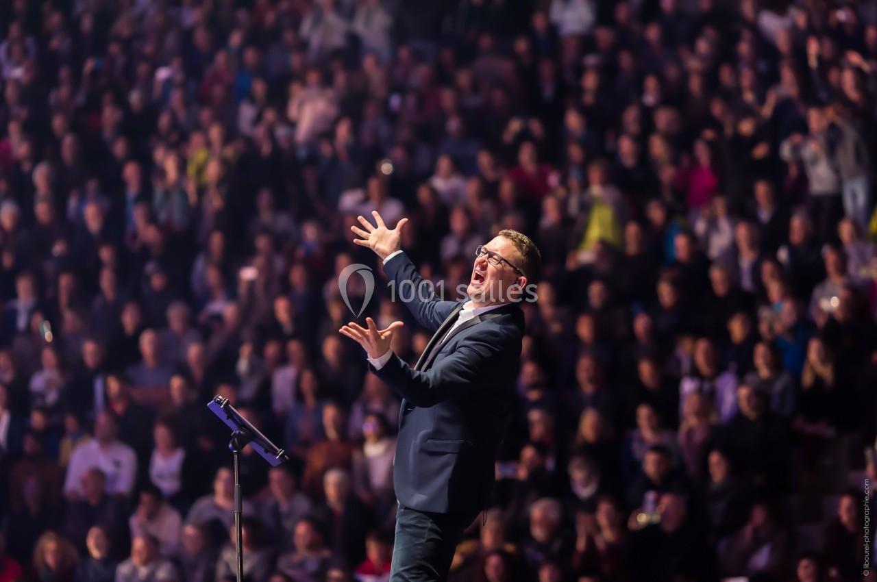 Un homme en costume gesticule avec énergie sur scène devant une foule dense et éclairée.