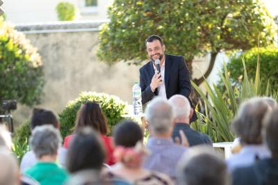 Une main verse du vin blanc dans un verre transparent, avec des bouteilles en arrière-plan flou.