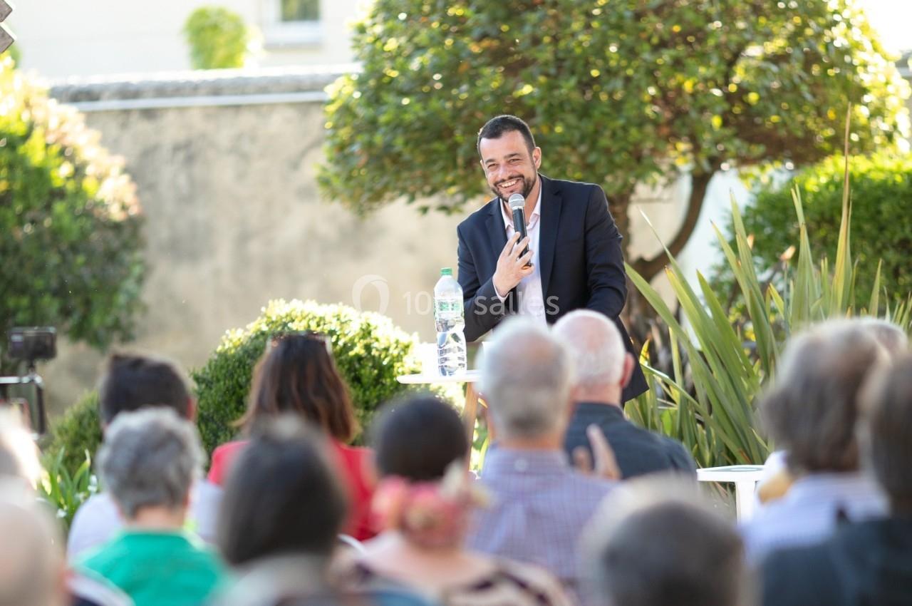 Un homme en costume sourit et parle devant un public en extérieur, avec des plantes et un mur en arrière-plan.