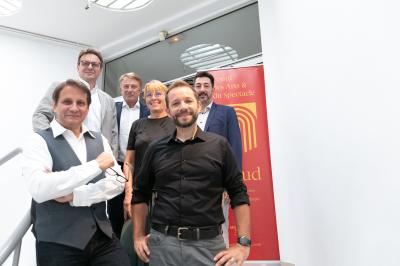 Une main verse du vin blanc dans un verre transparent, avec des bouteilles en arrière-plan flou.