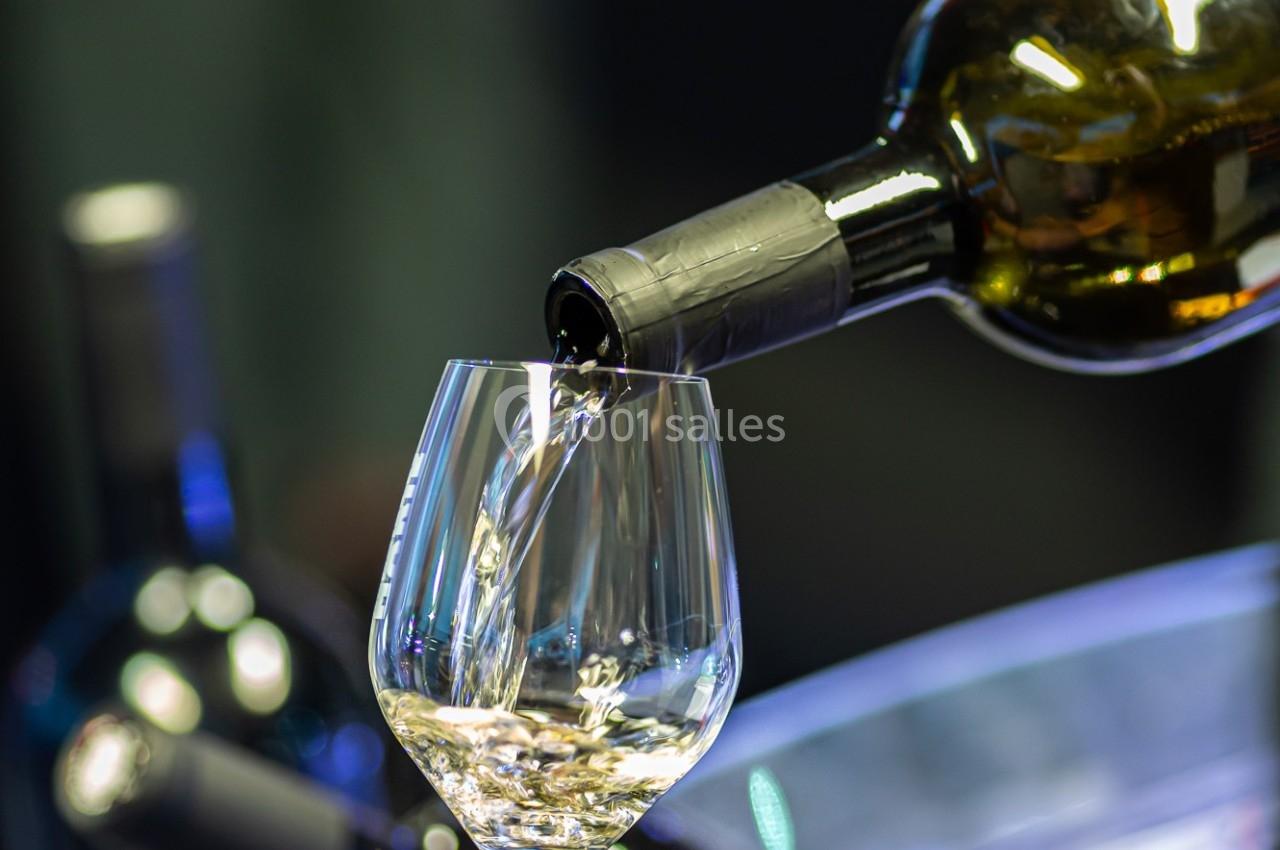 Une main verse du vin blanc dans un verre transparent, avec des bouteilles en arrière-plan flou.