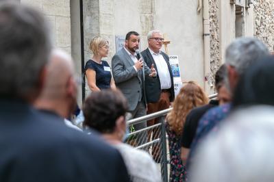 Une main verse du vin blanc dans un verre transparent, avec des bouteilles en arrière-plan flou.