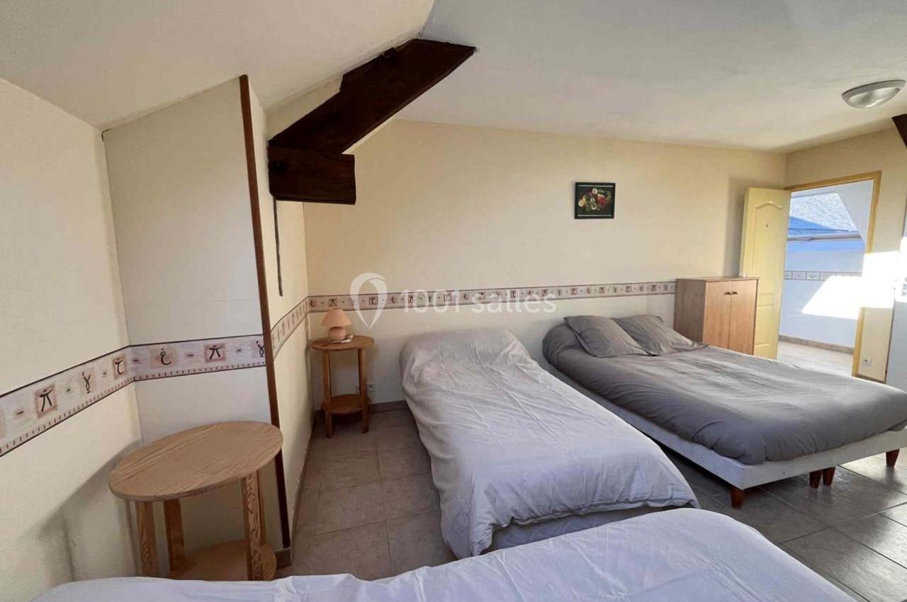 Chambre mansardée avec trois lits simples, murs clairs, mobilier en bois et lumière naturelle entrant par une fenêtre.