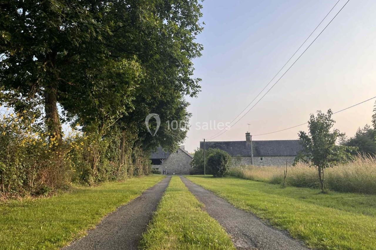 Allée bordée d'herbe menant à une maison en pierre entourée de champs et d'arbres sous un ciel clair.