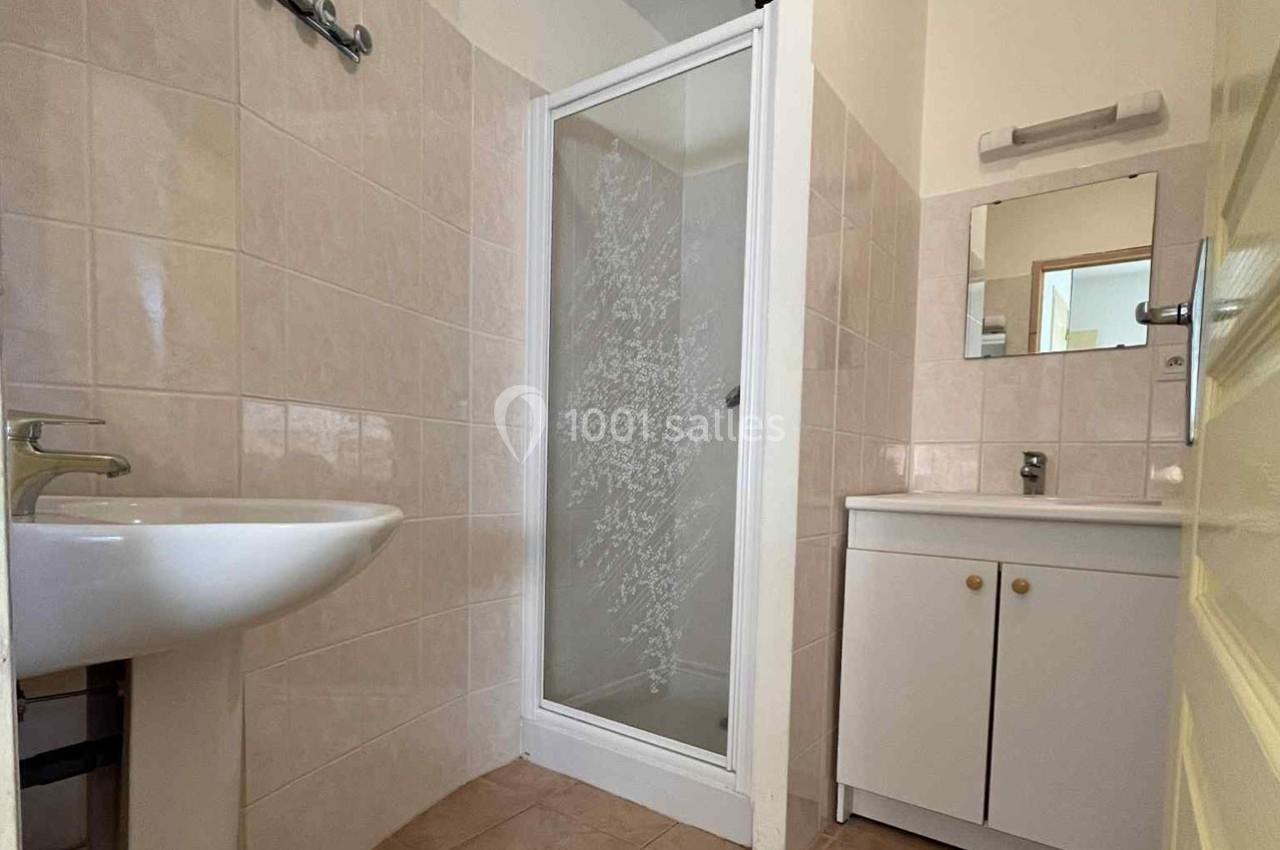 Salle de bain avec douche vitrée, lavabo mural, meuble sous vasque blanc et carrelage beige.