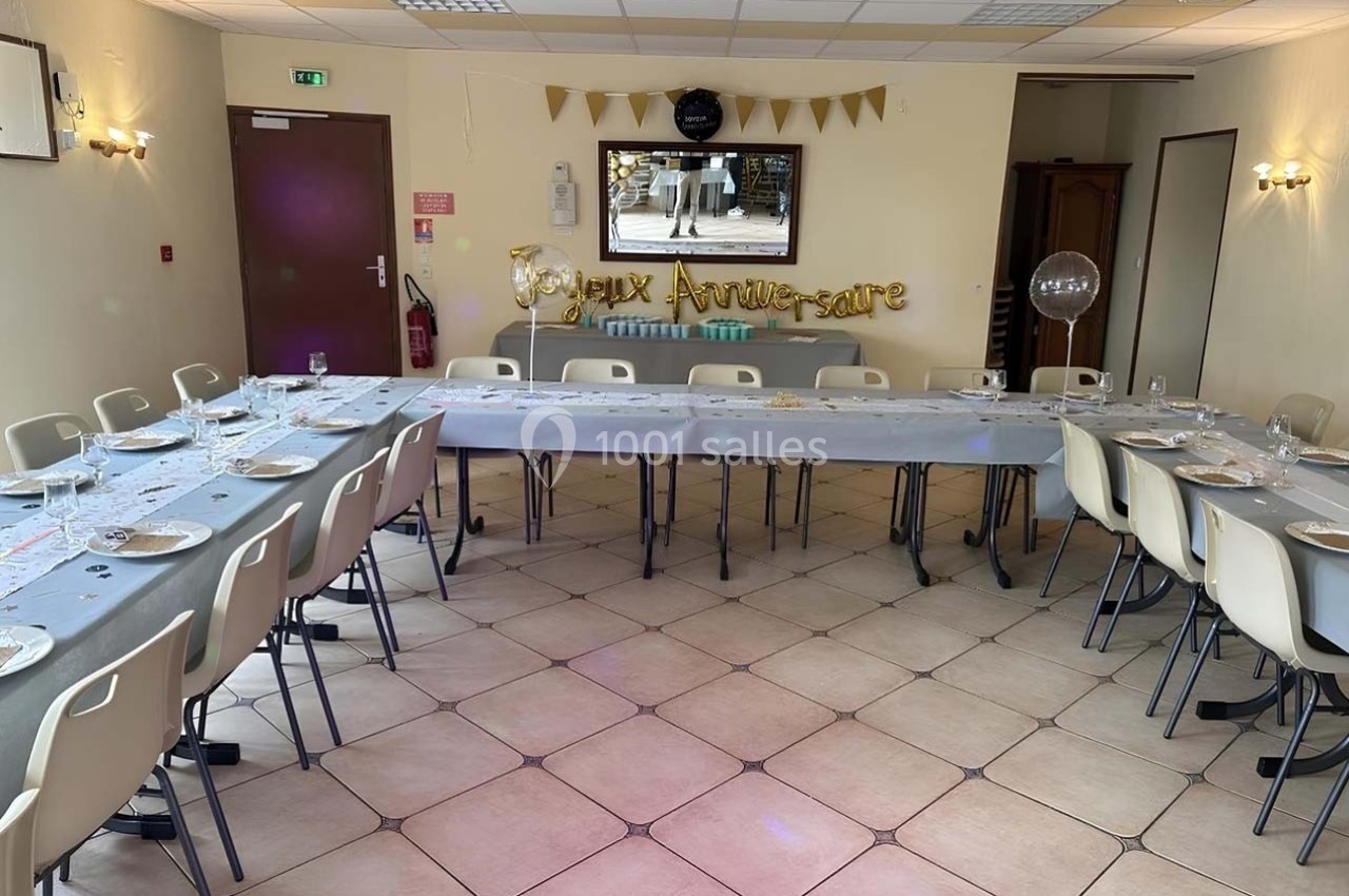 Salle décorée pour un anniversaire avec une grande table en U, nappes grises, vaisselle et guirlande ’Joyeux Anniversaire’.