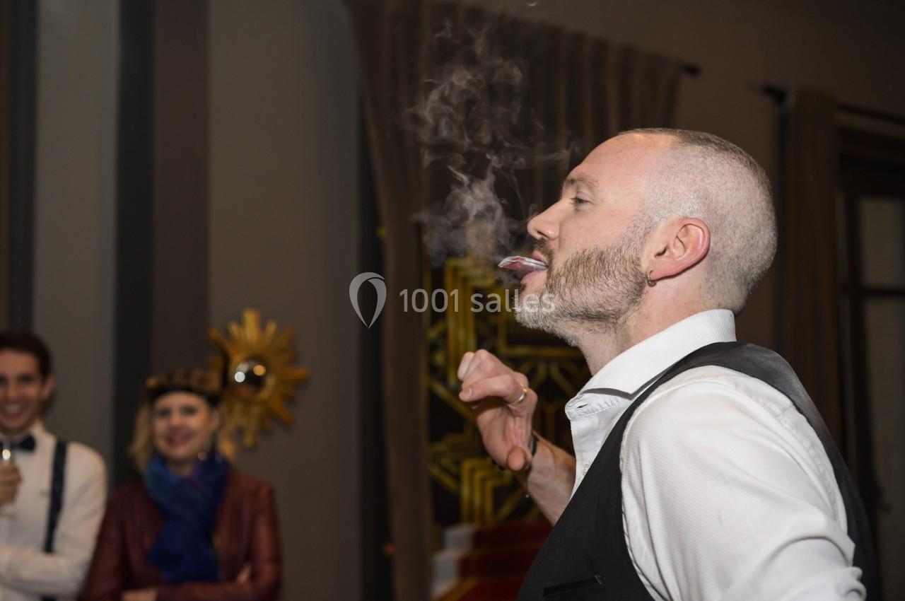 Un homme souffle de la fumée en l'air dans une pièce, avec des personnes en arrière-plan qui observent.