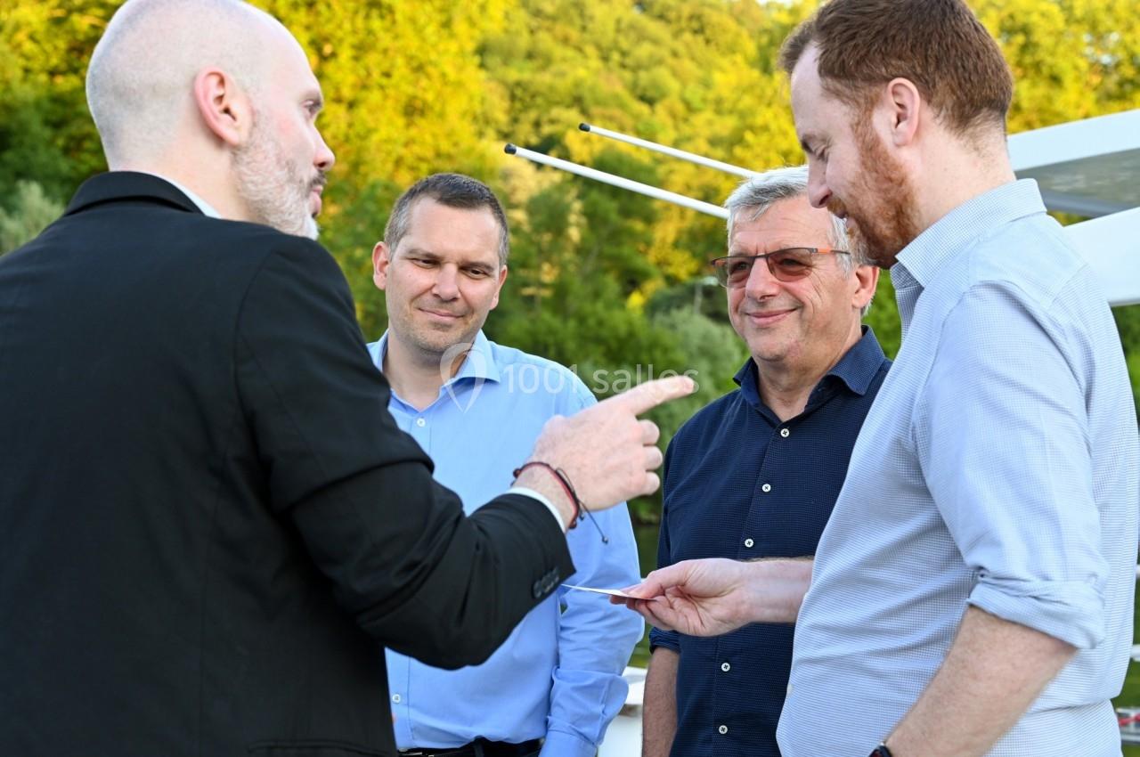 Quatre hommes discutent en extérieur, l'un tend une carte à un autre, avec des arbres en arrière-plan.
