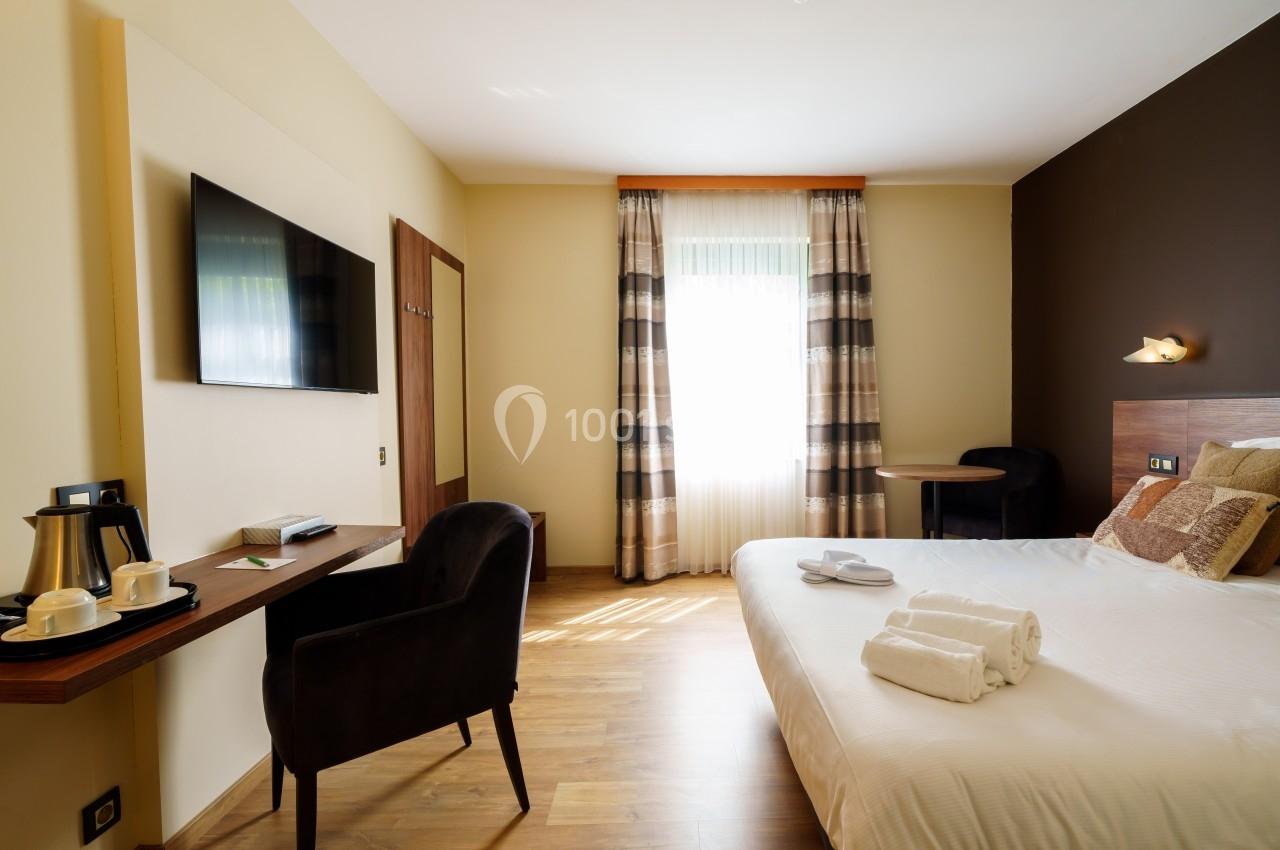 Chambre d'hôtel lumineuse avec lit double, bureau, télévision murale, fauteuil et fenêtre avec rideaux.