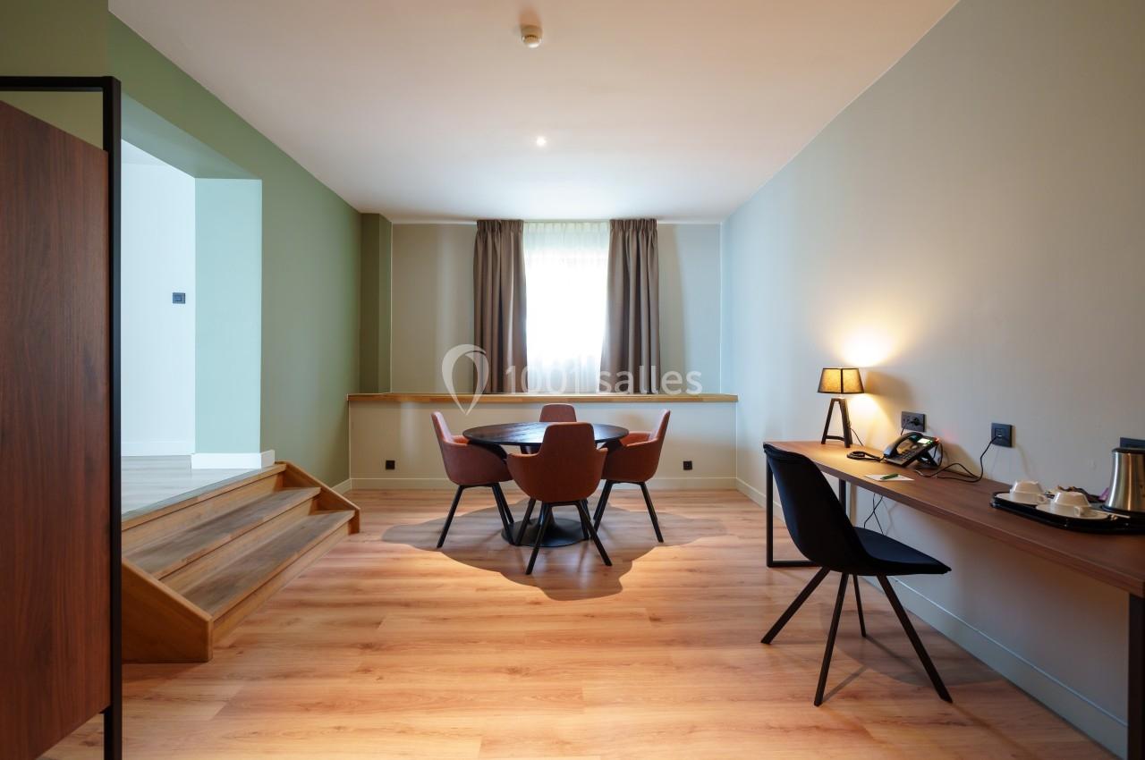 Salle lumineuse avec table ronde, chaises modernes, bureau équipé et sol en bois clair, éclairée par une fenêtre avec…