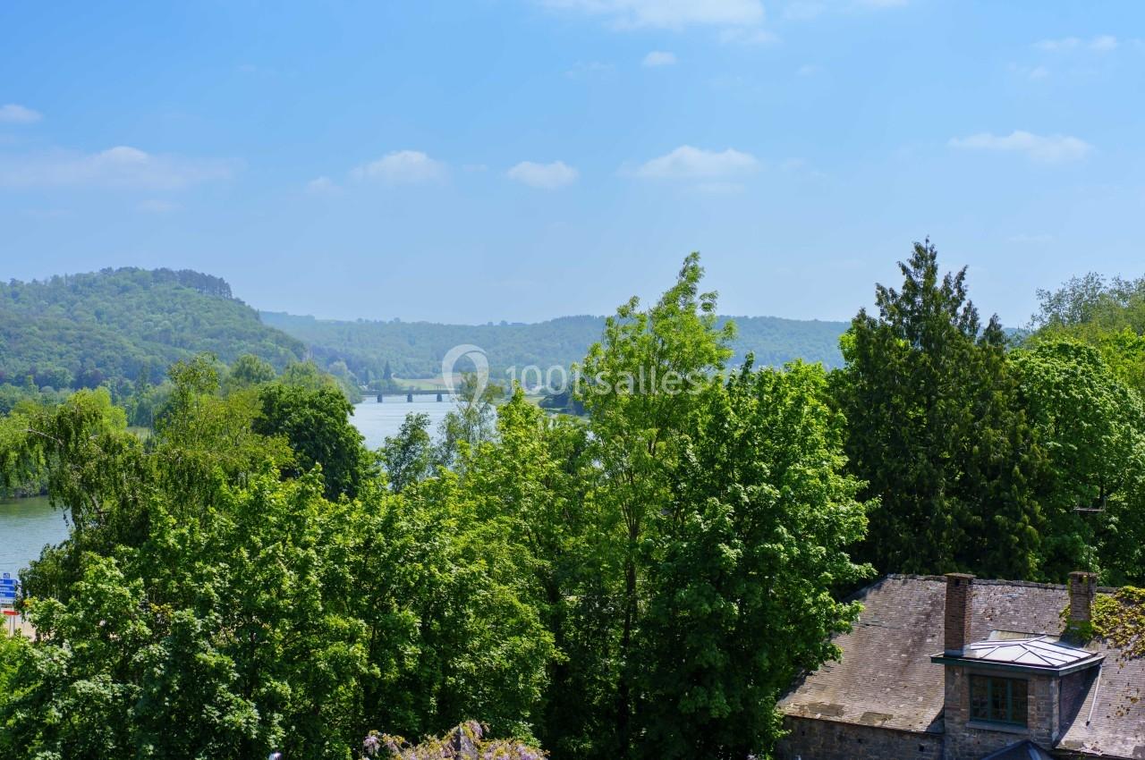 Vue sur une rivière entourée de collines boisées et de verdure, avec des toits de maisons au premier plan.