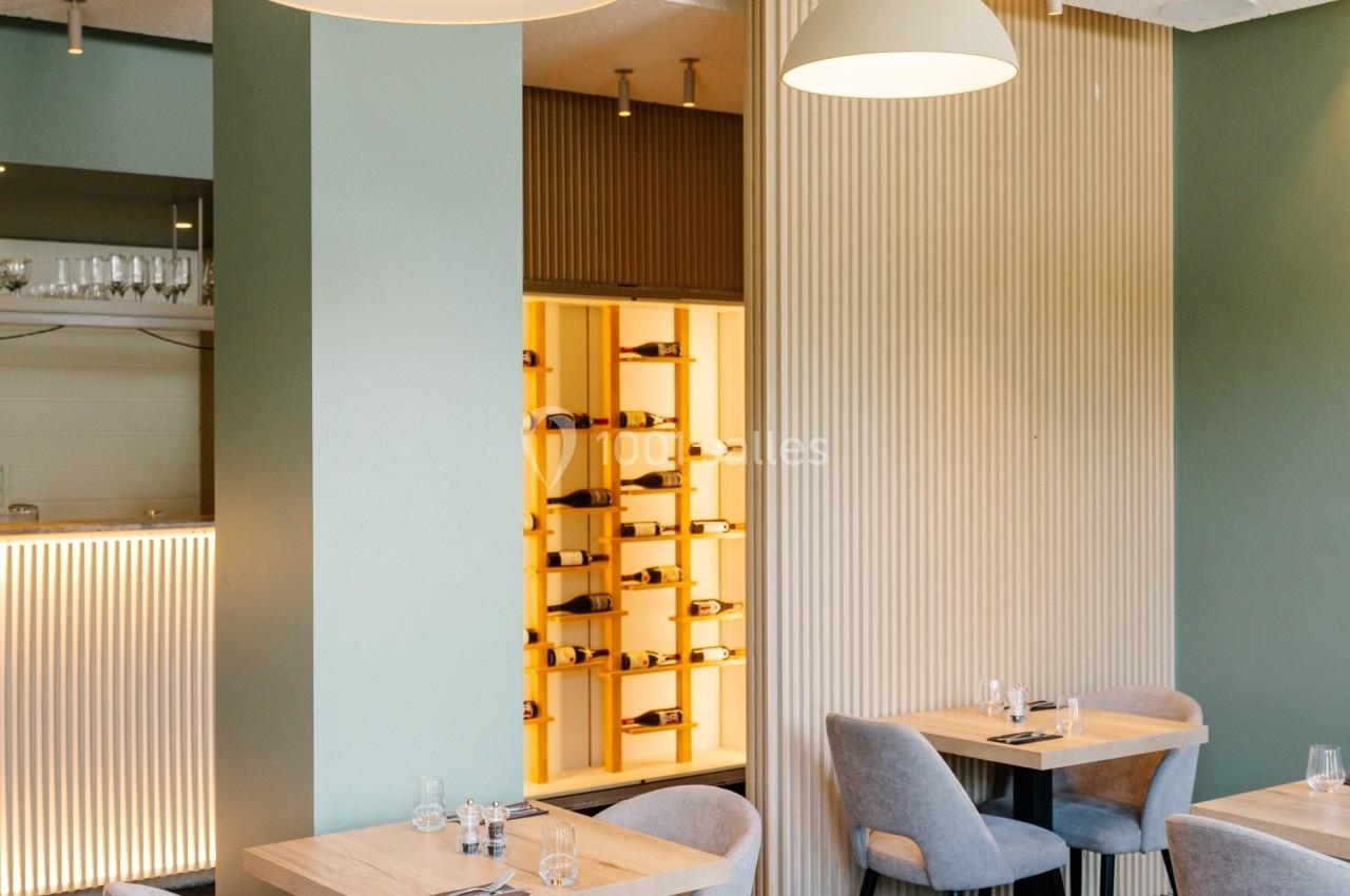 Salle de restaurant moderne avec tables en bois clair, chaises grises et éclairage suspendu, vue sur une cave à vin vitrée.