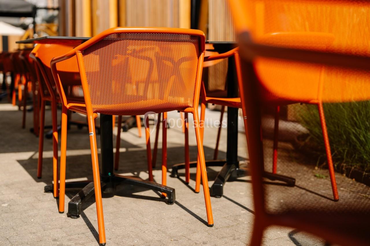 Chaises orange en métal disposées autour de tables sur une terrasse extérieure ensoleillée.