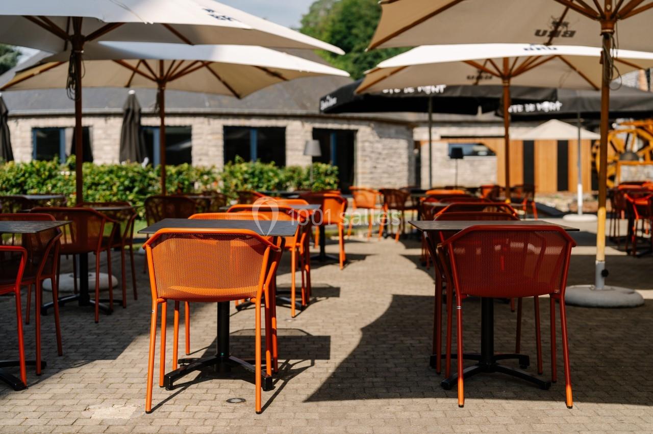 Terrasse extérieure avec tables et chaises orange sous des parasols blancs, entourée de verdure et de bâtiments en pierre.