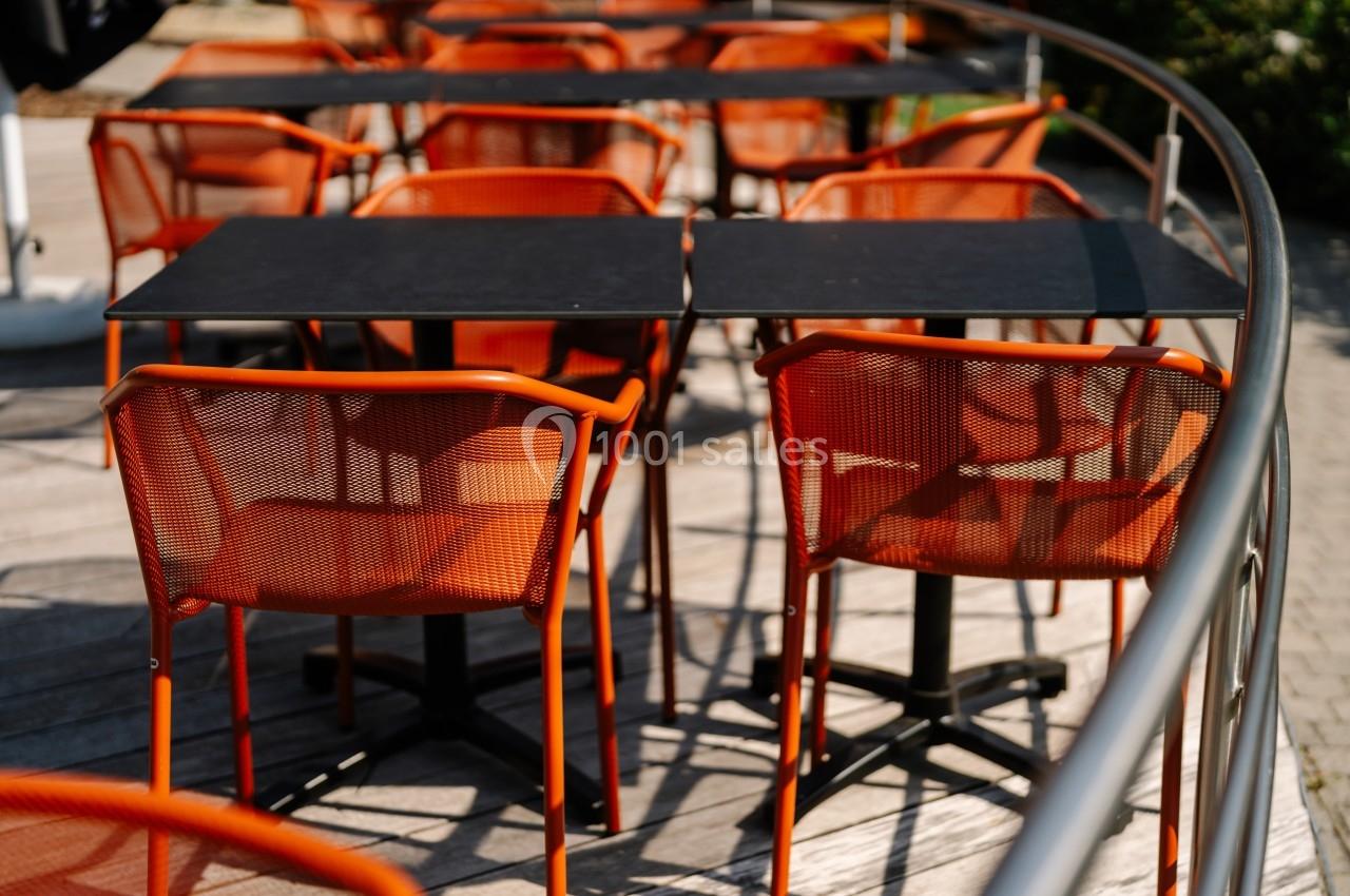 Chaises et tables en métal orange disposées sur une terrasse en bois, entourée d'une rambarde métallique.