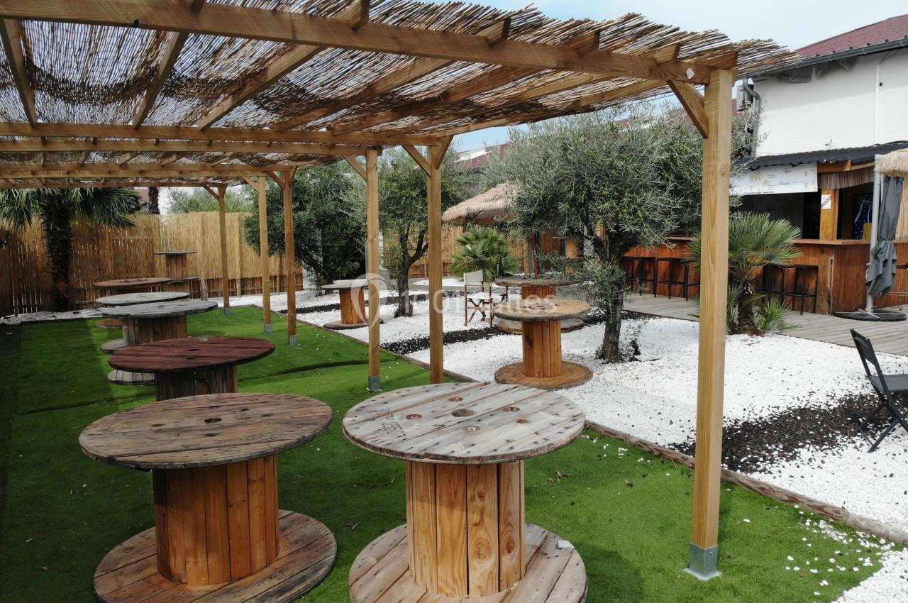 Terrasse extérieure avec des tables rondes en bois, pergola en bambou, sol en gazon synthétique et décor végétal.