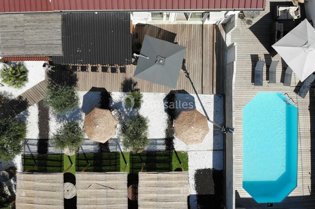 Vue aérienne d'une terrasse avec piscine, parasols, espaces verts, et zones aménagées en bois et gravier.