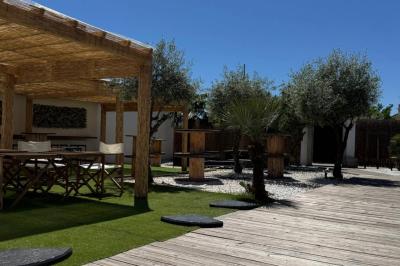 Terrasse extérieure avec pergola en bois, tables, chaises, oliviers et chemin en bois sur gazon et gravier.