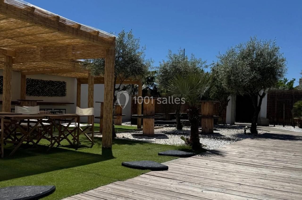 Terrasse extérieure avec pergola en bois, tables, chaises, oliviers et chemin en bois sur gazon et gravier.