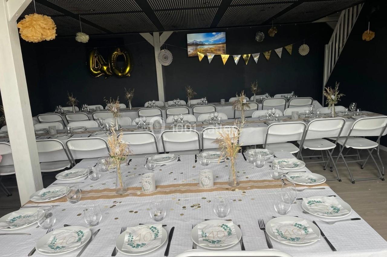 Salle décorée pour un événement, avec tables dressées, nappes blanches, vaisselle élégante et guirlandes suspendues.